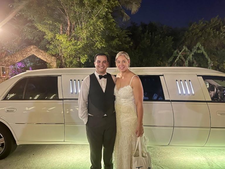 A couple in their wedding outfits standing outside of a white limo.